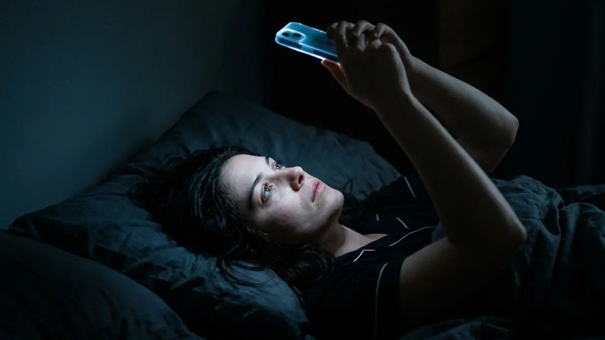 A woman lying in a dark bedroom at night, her face illuminated by the blue-white glow of her phone screen, representing relationship addiction loops