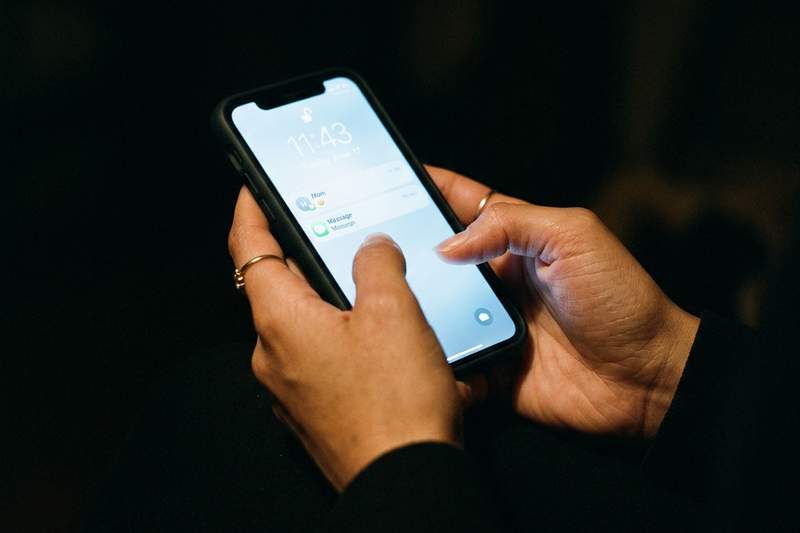Close-up of a woman's hands holding a phone in near darkness, the screen's cool blue-white glow illuminating her palms and fingers